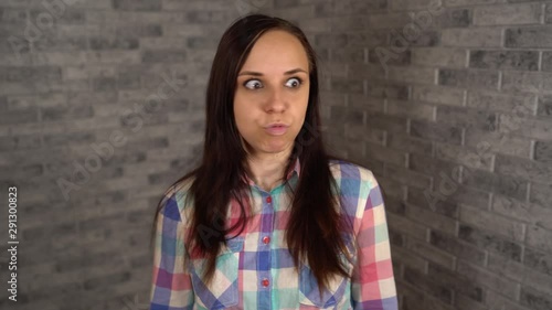 A beautiful young woman in a plaid shirt resents and expresses her negative emotions on a brick background.