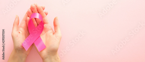 Breast cancer awareness campaign horizontal banner with copy space. Woman   s hands holding pink breast cancer awareness ribbon over pink background. Healthcare and medicine concept.