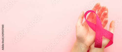 Breast cancer awareness month horizontal banner with copy space. Woman’s hand holding pink ribbon over pink background. Healthcare and medicine concept.