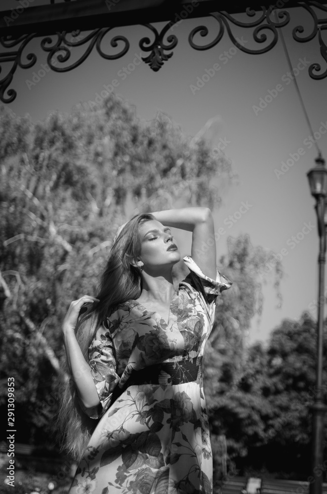 A young and beautiful girl with professional makeup in a beautiful summer dress stands in the sun in a city park. Summer black and white photo.