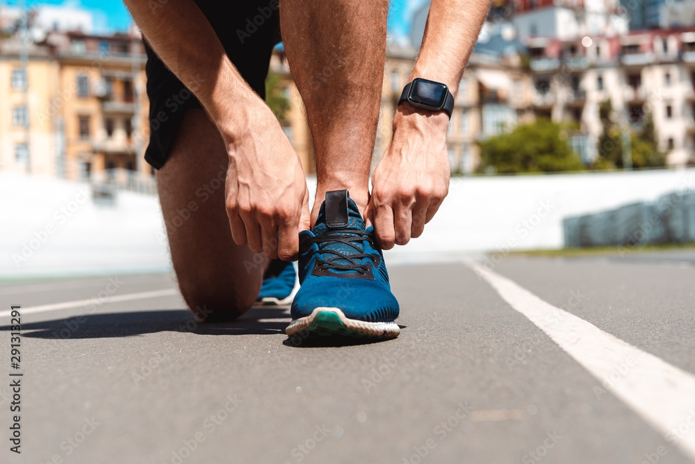 cropped view of athletic young sportsman in smartwatch touching sneakers on running track with buildings on background