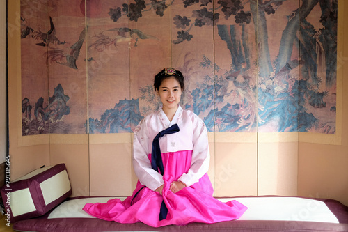 Korean girl sit on mat,place her hands on her lap,sitting on the mat,wearing hanbok dress in Korean traditional pink color so beautiful with her smile,she take a seat in front old drawing. 