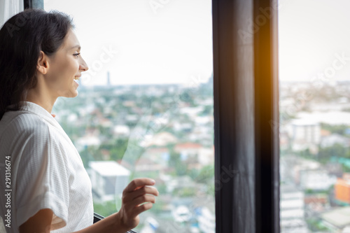 Beautiful woman look at nice view of cityscape at condominium room or hotel of hig building. Gorgeous woman wear bathrobe. Pretty girl get fresh, happiness with smiley face. Glamour lady feel relaxing