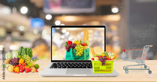 supermarket aisle blurred background with laptop computer and shopping cart on wood table grocery online concept