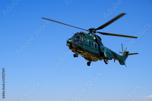 Big dark green hovering military helicopter on blue sky background