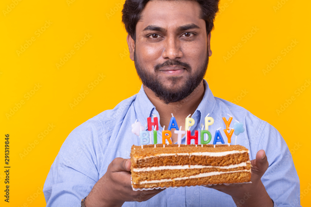 Close-up of crazy cheerful young indian man in paper congratulatory hat holding cakes happy birthday standing on a yellow background. Jubilee congratulations concept. Close-up of crazy cheerful young indian man in paper congratulatory hat holding cakes happy birthday standing on a yellow background. Jubilee congratulations concept.