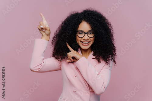 Half length shot of cheerful Afro American woman with dark bushy hairstyle, wears violet suit, points above with both index fingers, demonstrates way to something, invites you going upstairs photo