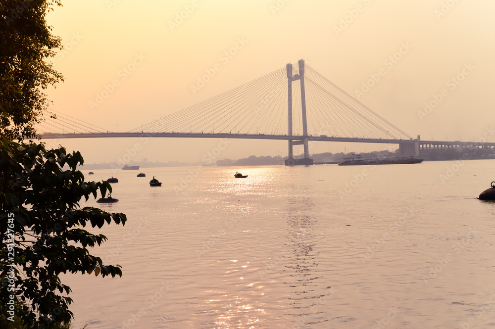 Obraz premium Sunset over Vidyasagar Setu (a modern Cantilever Spar Cable-Stayed Bridge) in a summer evening on the river Hooghly. Silhouette view from distant. Howrah city, Kolkata, West Bengal, India, South Asia.
