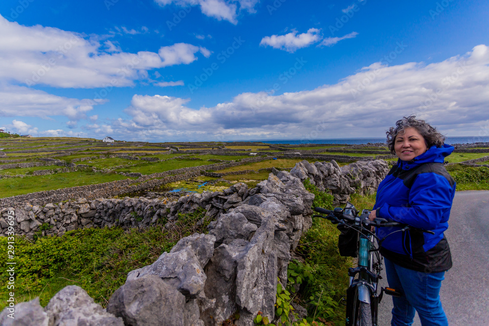 Beautiful and smiling female cyclist pausing on a road next to a stone fence on the Inis Oirr island, field with limestone fences and the sea in the background, wonderful day in Aran Islands, Ireland