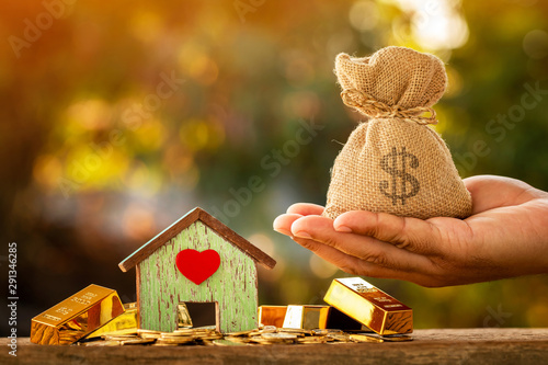 Man hand hold money bag with presenting and home model put on the gold bar and coin with growing interest in the public park, Saving money for buy house or loan for investment of real estate concept. photo