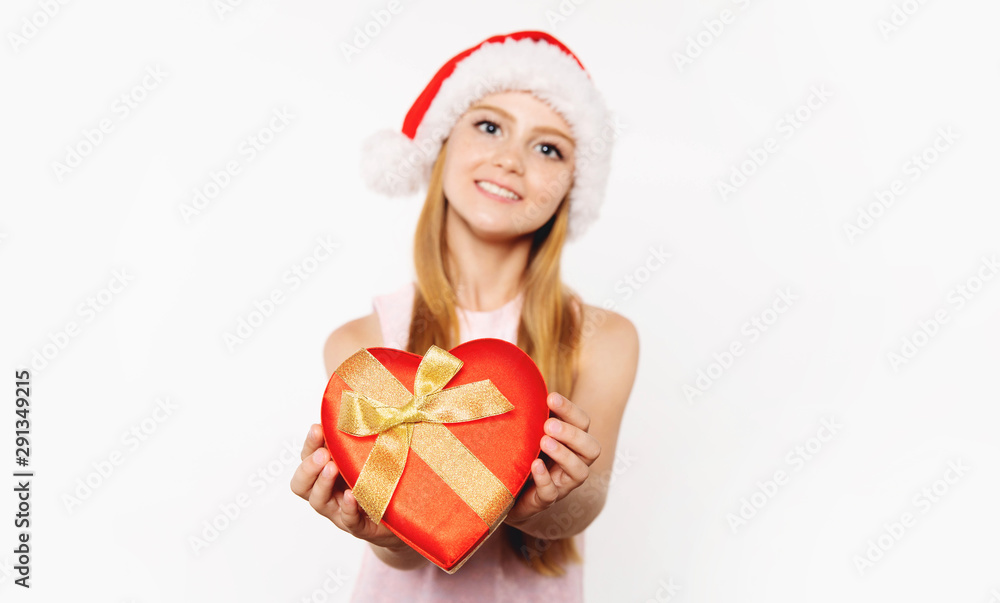 Fototapeta premium With love from me to you. Cute teenage blonde and freckled girl poses with genuine smile while wearing Santa hat and offering a red heart-shaped box with a golden ribbon
