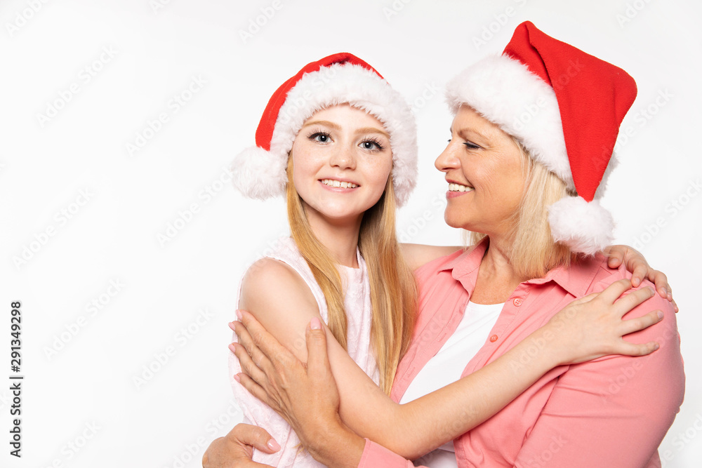 Beautiful senior grand mother in a pink shirt and Santa hat with light shoulder-length hair hugs her teenage blonde grand daughter in Christmas hat and laughs with her while looking at the camera.
