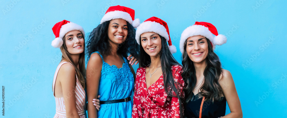Enjoying our youth. Close-up of a group of four dark-haired girls in Santa caps posing and smiling for the joint photo over blue background Enjoying our youth. Close-up of a group of four dark-haired girls in Santa caps posing and smiling for the joint photo over blue background