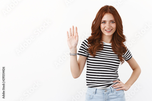 Cute friendly and casual young redhead caucasian girl in striped t-shirt, waving raised hand in greeting gesture, smiling joyfully, welcome friend, invite guest, saying hello or hi, white background