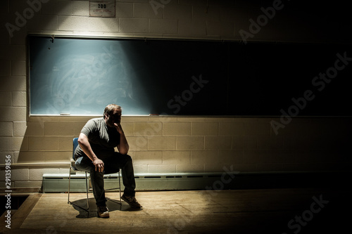 Caucasian Man Sitting in Thought on an Empty School Chair in a Dark, Shadowy Classroom - in Front of a Chalkboard with a Single Beam of Light Overhead