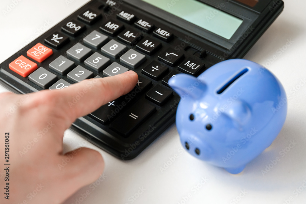Close up of finger presses the calculator button near piggy bank.