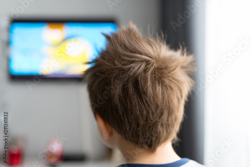 A child of several years standing back in the room looking at the television.