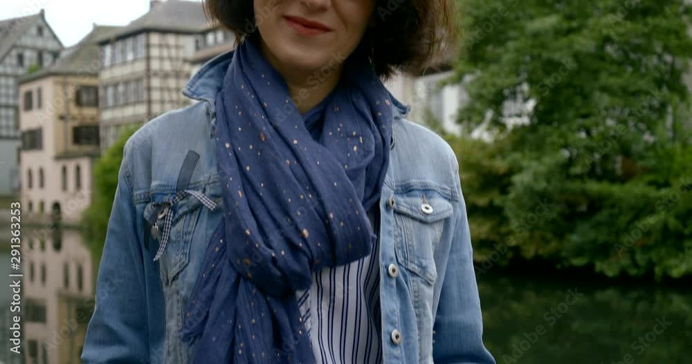 Portrait of a dark-haired fashionable smiling woman in middle-aged glasses, she is set against a backdrop of small beautiful European houses, bushes and a river. Portrait of a dark-haired fashionable smiling woman in middle-aged glasses, she is set against a backdrop of small beautiful European houses, bushes and a river.