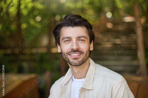 Close-up of beautiful bearded male with dark hair posing over green ciy park on sunny day, looking to camera with charming and sincere smile