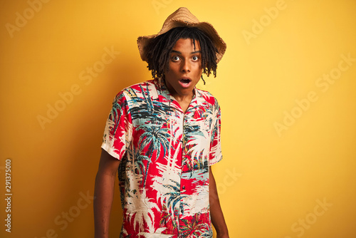 Afro american man with dreadlocks wearing floral shirt and hat over isolated yellow background afraid and shocked with surprise expression, fear and excited face.