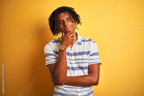 Afro american man with dreadlocks wearing striped shirt over isolated yellow background looking confident at the camera with smile with crossed arms and hand raised on chin. Thinking positive. © Krakenimages.com