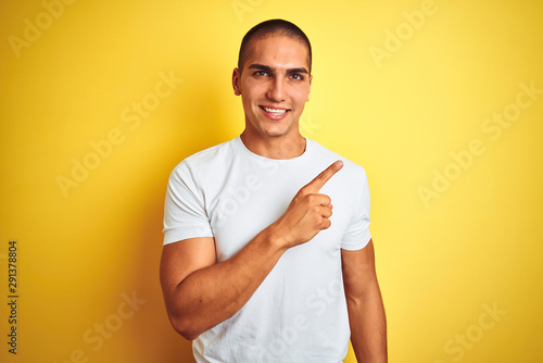 Young caucasian man wearing casual white t-shirt over yellow isolated background cheerful with a smile of face pointing with hand and finger up to the side with happy and natural expression on face