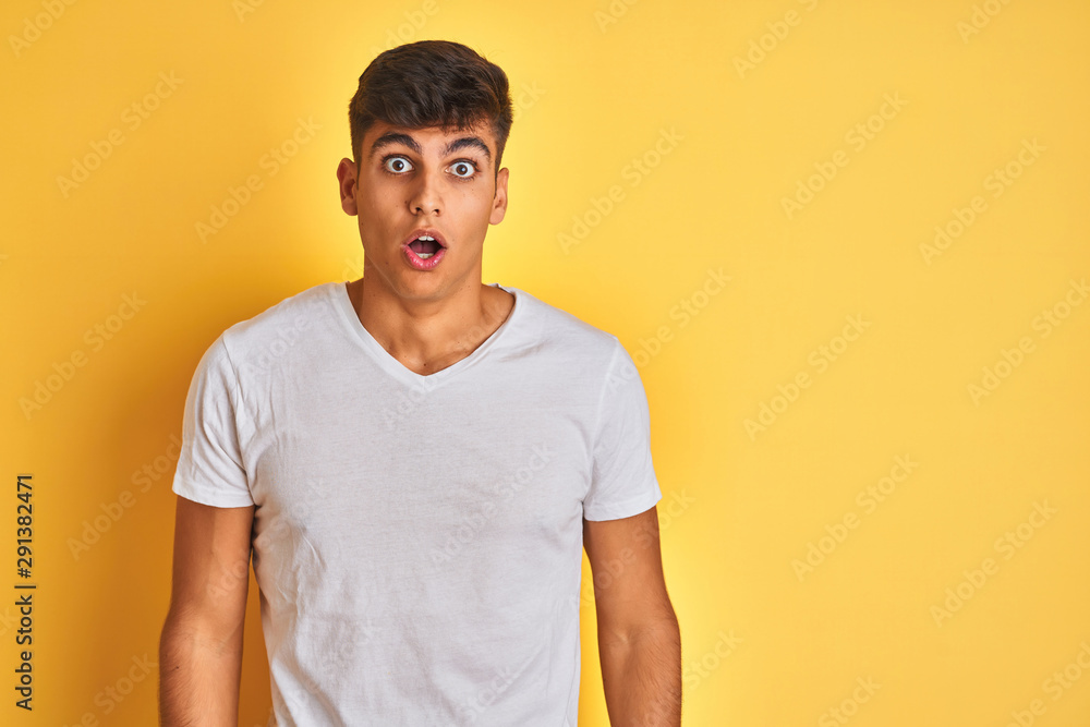 Young indian man wearing white t-shirt standing over isolated yellow background afraid and shocked with surprise and amazed expression, fear and excited face. Young indian man wearing white t-shirt standing over isolated yellow background afraid and shocked with surprise and amazed expression, fear and excited face.