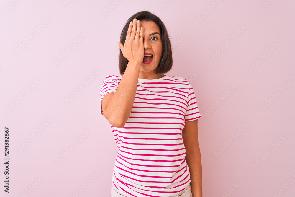 Young beautiful woman wearing striped t-shirt standing over isolated pink background covering one eye with hand, confident smile on face and surprise emotion. Young beautiful woman wearing striped t-shirt standing over isolated pink background covering one eye with hand, confident smile on face and surprise emotion.