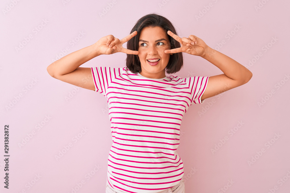 Young beautiful woman wearing striped t-shirt standing over isolated pink background Doing peace symbol with fingers over face, smiling cheerful showing victory Young beautiful woman wearing striped t-shirt standing over isolated pink background Doing peace symbol with fingers over face, smiling cheerful showing victory