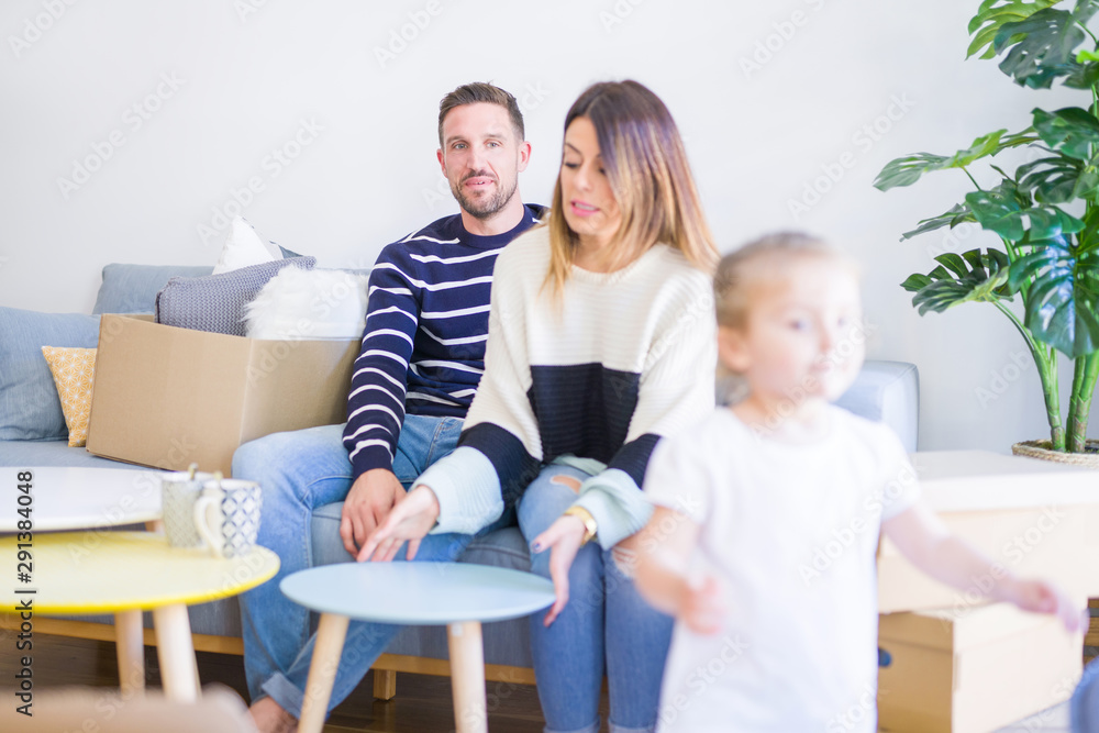 Beautiful family, parents sitting on the sofa drinking coffee looking his kid playing at new home around cardboard boxes