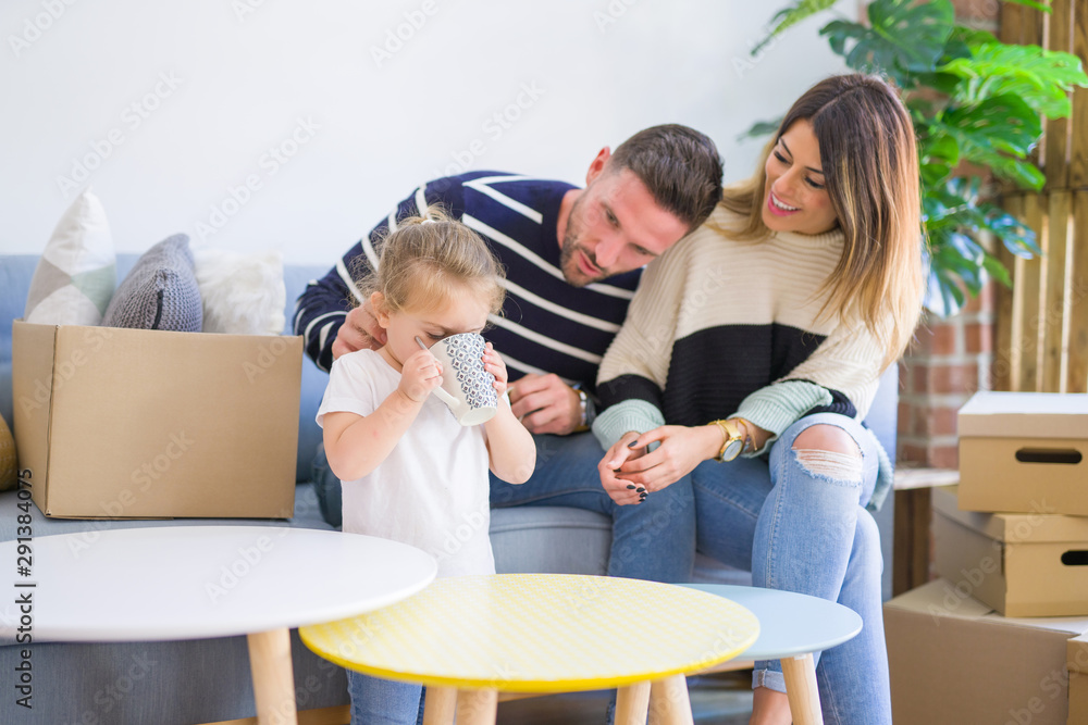 Beautiful family, parents sitting on the sofa drinking coffee looking his kid playing at new home around cardboard boxes Beautiful family, parents sitting on the sofa drinking coffee looking his kid playing at new home around cardboard boxes