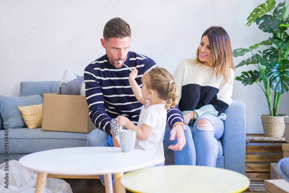 Beautiful family, parents sitting on the sofa drinking coffee looking his kid playing at new home around cardboard boxes Beautiful family, parents sitting on the sofa drinking coffee looking his kid playing at new home around cardboard boxes