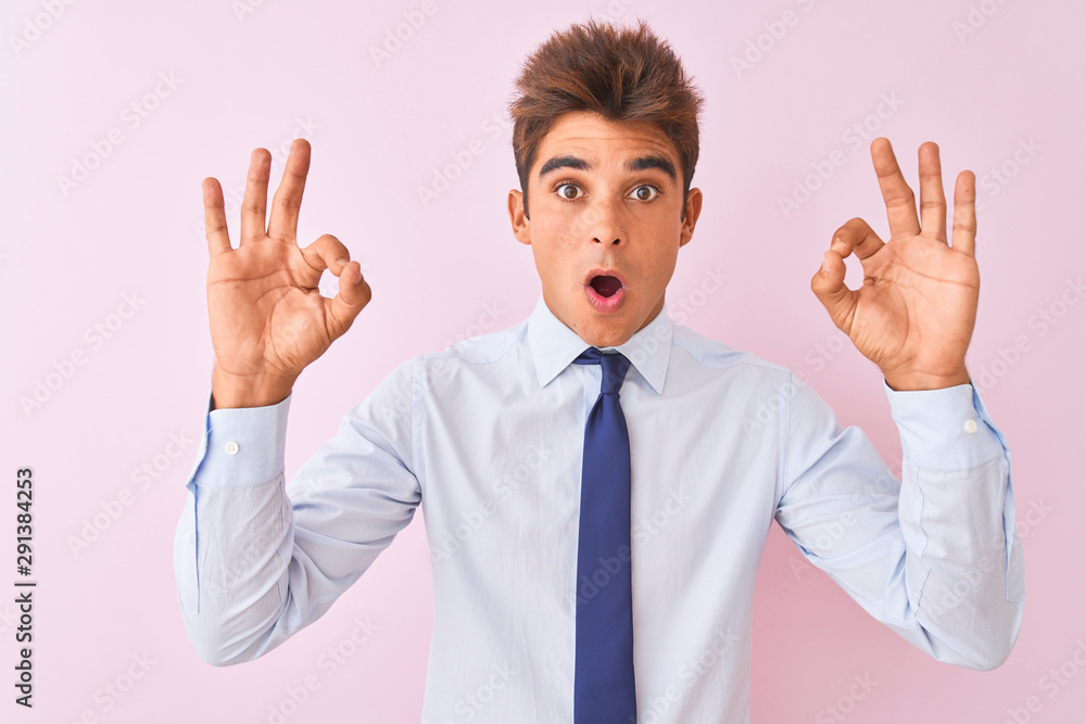 Young handsome businessman wearing shirt and tie standing over isolated pink background looking surprised and shocked doing ok approval symbol with fingers. Crazy expression