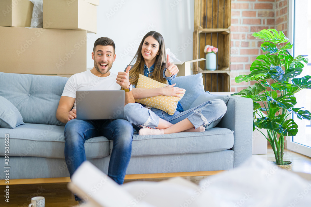 Young beautiful couple relaxing sitting on the sofa of new house using computer laptop happy with big smile doing ok sign, thumb up with fingers, excellent sign Young beautiful couple relaxing sitting on the sofa of new house using computer laptop happy with big smile doing ok sign, thumb up with fingers, excellent sign