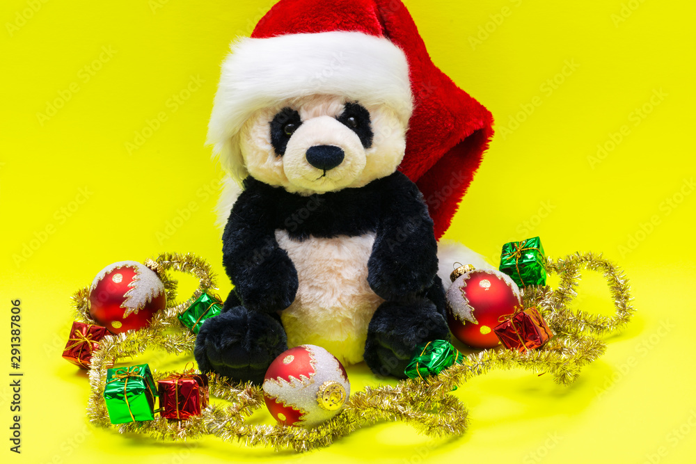 Panda Bear plush toy with Red Christmas Santa Hat surrounded by Tinsel Garland and Vintage Ornaments on bright yellow background. Holidays concept. Panda Bear plush toy with Red Christmas Santa Hat surrounded by Tinsel Garland and Vintage Ornaments on bright yellow background. Holidays concept.