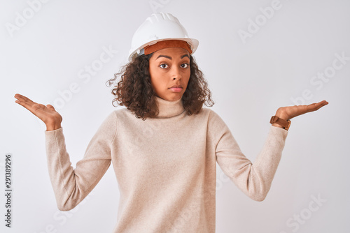 Young brazilian architect woman wearing security helmet over isolated white background clueless and confused expression with arms and hands raised. Doubt concept.
