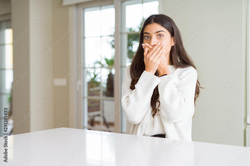 Young beautiful woman at home on white table shocked covering mouth with hands for mistake. Secret concept. Young beautiful woman at home on white table shocked covering mouth with hands for mistake. Secret concept.