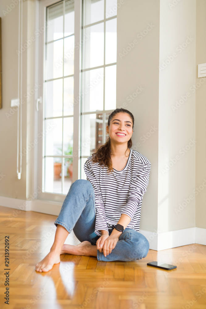 Young beautiful woman sitting on the floor at home looking away to side with smile on face, natural expression. Laughing confident. Young beautiful woman sitting on the floor at home looking away to side with smile on face, natural expression. Laughing confident.