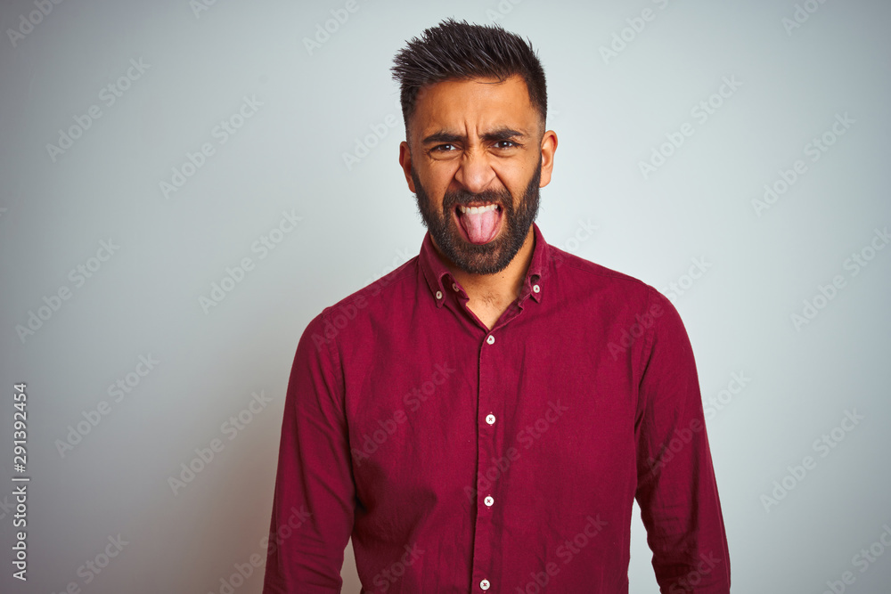 Obraz premium Young indian man wearing red elegant shirt standing over isolated grey background sticking tongue out happy with funny expression. Emotion concept.