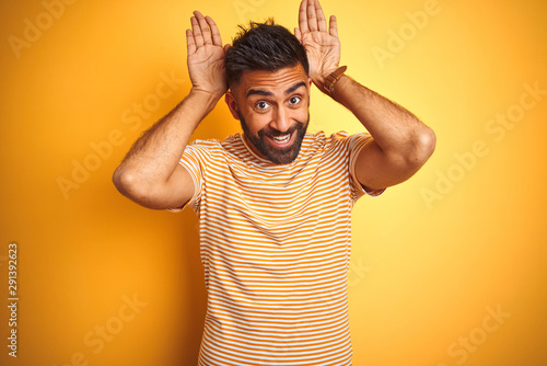 Young indian man wearing t-shirt standing over isolated yellow background Doing bunny ears gesture with hands palms looking cynical and skeptical. Easter rabbit concept.