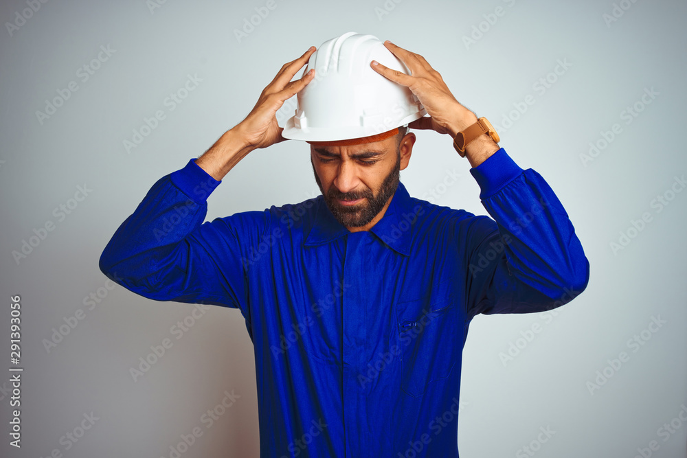 Handsome indian worker man wearing uniform and helmet over isolated white background suffering from headache desperate and stressed because pain and migraine. Hands on head. Handsome indian worker man wearing uniform and helmet over isolated white background suffering from headache desperate and stressed because pain and migraine. Hands on head.