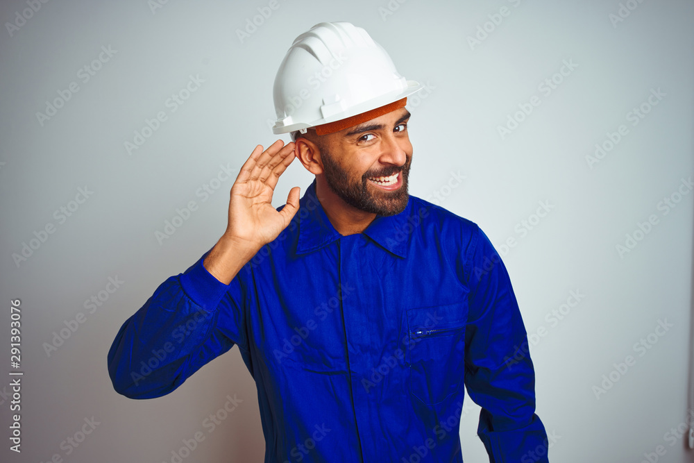 Handsome indian worker man wearing uniform and helmet over isolated white background smiling with hand over ear listening an hearing to rumor or gossip. Deafness concept. Handsome indian worker man wearing uniform and helmet over isolated white background smiling with hand over ear listening an hearing to rumor or gossip. Deafness concept.