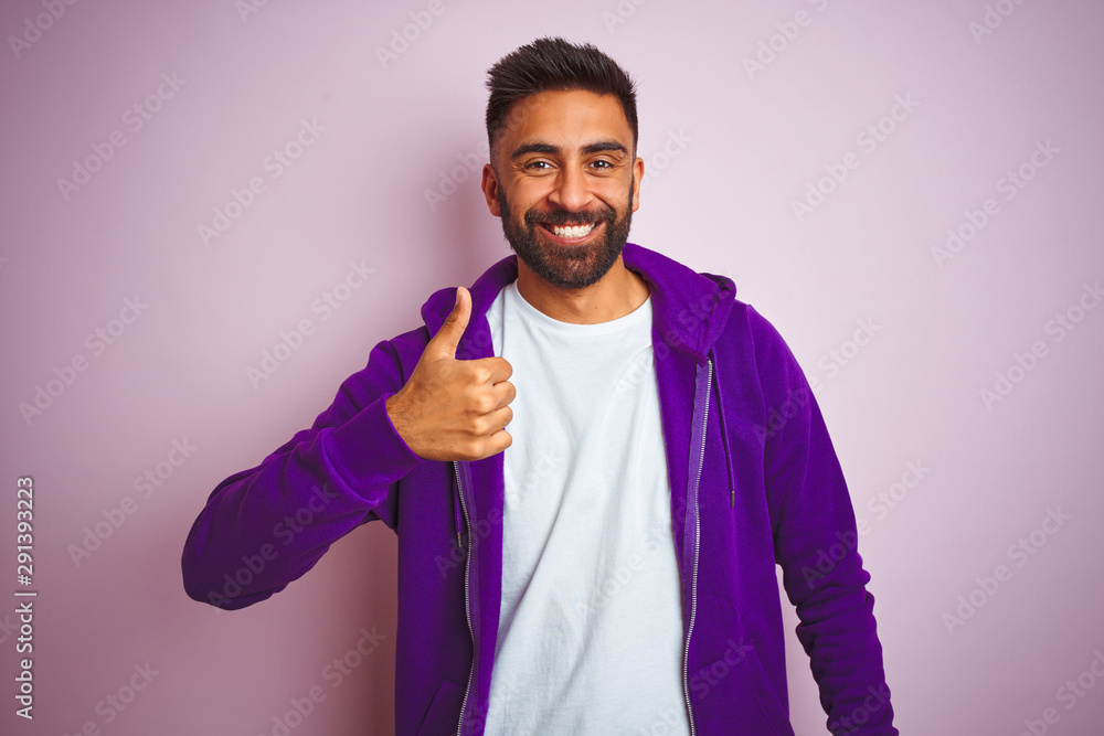 Young indian man wearing purple sweatshirt standing over isolated pink background doing happy thumbs up gesture with hand. Approving expression looking at the camera showing success. Young indian man wearing purple sweatshirt standing over isolated pink background doing happy thumbs up gesture with hand. Approving expression looking at the camera showing success.