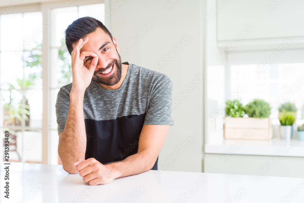 Handsome hispanic man wearing casual t-shirt at home doing ok gesture with hand smiling, eye looking through fingers with happy face. Handsome hispanic man wearing casual t-shirt at home doing ok gesture with hand smiling, eye looking through fingers with happy face.