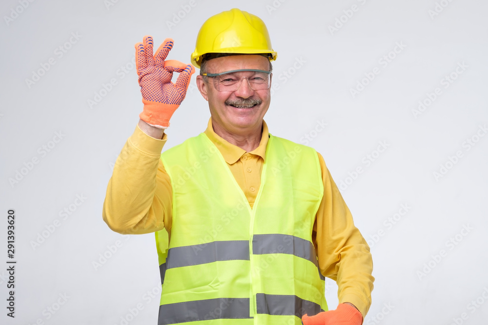 Senior hispanic construction worker in hard hat showing ok sign smiling on gray background