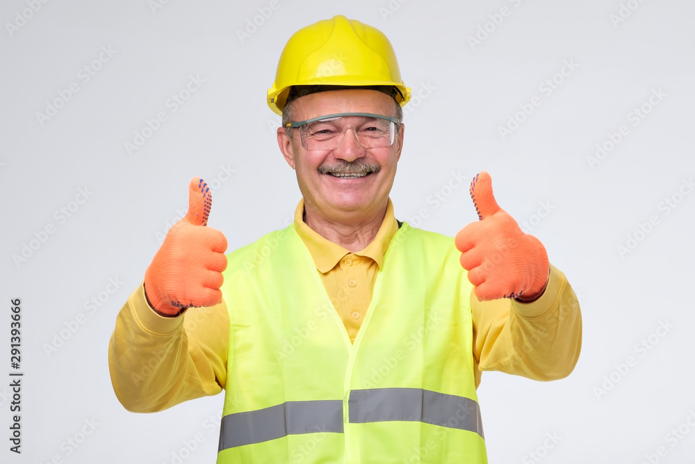 Senior hispanic construction worker in hard hat showing thumb up sign smiling on gray background