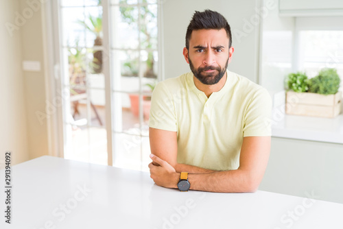 Handsome hispanic man casual yellow t-shirt at home skeptic and nervous, disapproving expression on face with crossed arms. Negative person.
