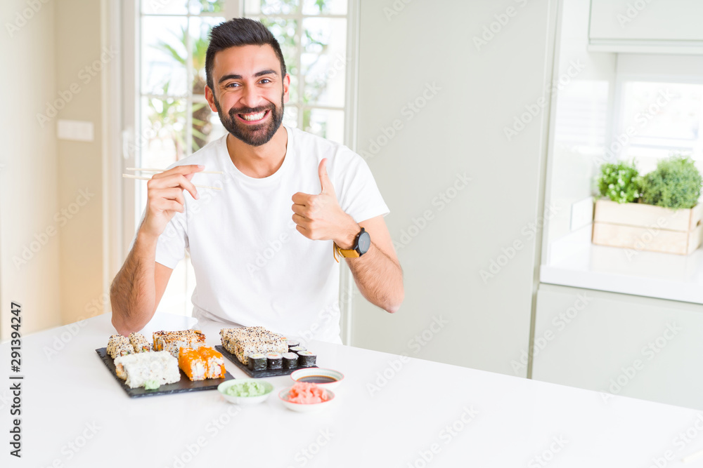 Handsome hispanic man eating asian sushi using chopsticks happy with big smile doing ok sign, thumb up with fingers, excellent sign Handsome hispanic man eating asian sushi using chopsticks happy with big smile doing ok sign, thumb up with fingers, excellent sign