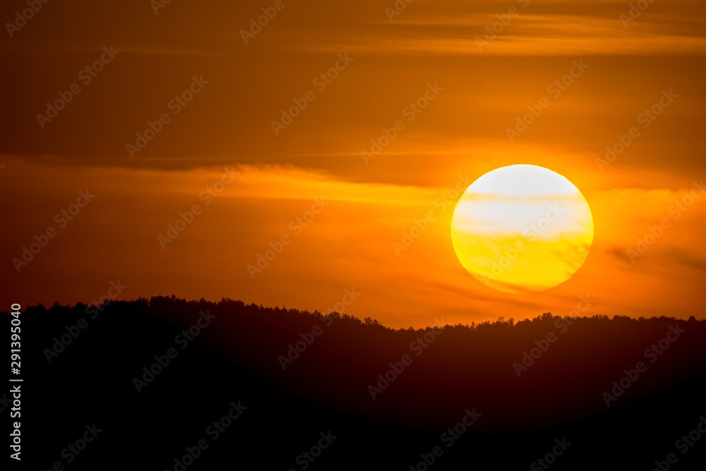 view of the big orange circle of the sun during sunset in the evening against the background of forest on hills and Cirrus clouds
