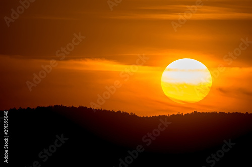 view of the big orange circle of the sun during sunset in the evening against the background of forest on hills and Cirrus clouds